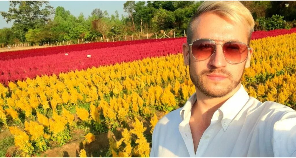 Casey Pownall faces the camera, wearing sunglasses on a sunny day, the backdrop is a field of rows of colourful flowers in red and yellow